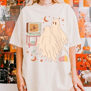Comfort Colors 1717 Shirt
Color: Ivory (off white cream)
Graphic: Two cartoon-style ghosts watch a witch flying on a TV screen surrounded by stars, popcorn, VHS tapes, and crescent moons.