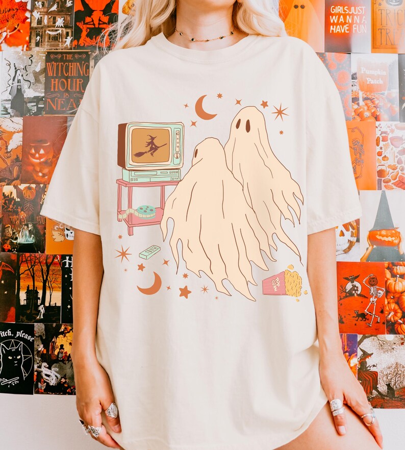 Comfort Colors 1717 Shirt
Color: Ivory (off white cream)
Graphic: Two cartoon-style ghosts watch a witch flying on a TV screen surrounded by stars, popcorn, VHS tapes, and crescent moons.