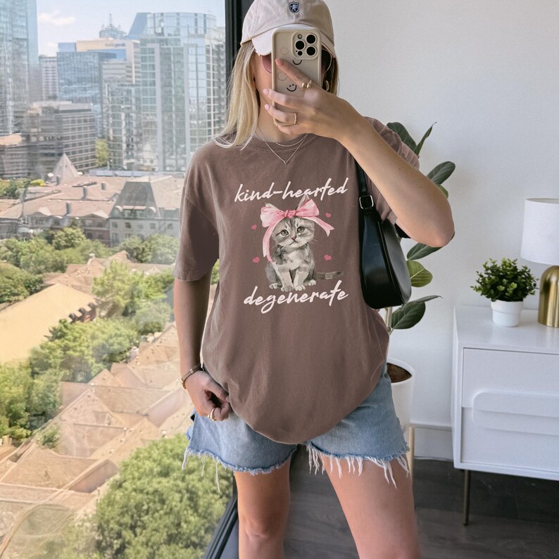 Comfort Colors 1717 Shirt
Color: Espresso Brown
Graphic: Illustration of a gray tabby kitten sitting with a large pink bow tied on its head. The kitten is surrounded by small pink hearts and framed by the handwritten-style phrase "kind-hearted degenerate" in soft, playful lettering.