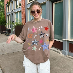 Comfort Colors 1717 T- Shirt
Color: Espresso (brown)
Graphic: bold star graphic design in multiple colors and textures. Features a mix of embroidered, metallic, and printed stars, along with abstract celestial details.