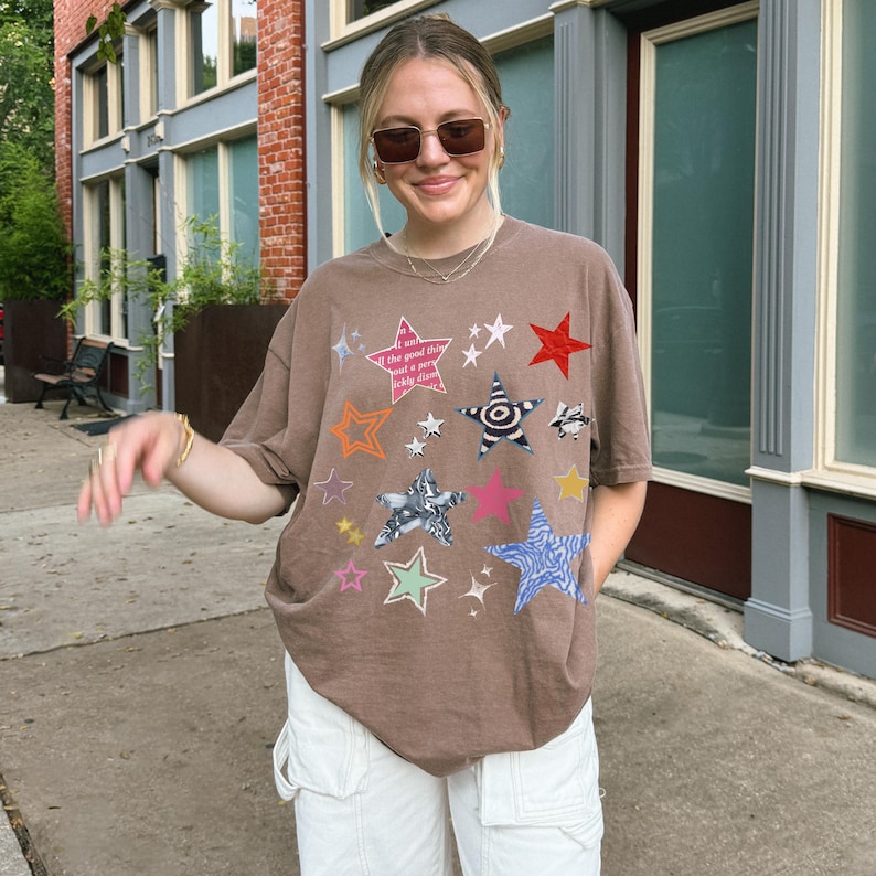 Comfort Colors 1717 T- Shirt
Color: Espresso (brown)
Graphic: bold star graphic design in multiple colors and textures. Features a mix of embroidered, metallic, and printed stars, along with abstract celestial details.