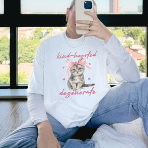 Comfort Colors 1717 Shirt
Color: White
Graphic: Illustration of a gray tabby kitten sitting with a large pink bow tied on its head. The kitten is surrounded by small pink hearts and framed by the handwritten-style phrase "kind-hearted degenerate" in soft, playful lettering.