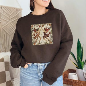 May include: A brown sweatshirt with a vintage-style illustration of two fairies dancing in a floral frame. The fairies are wearing colorful dresses and have wings.