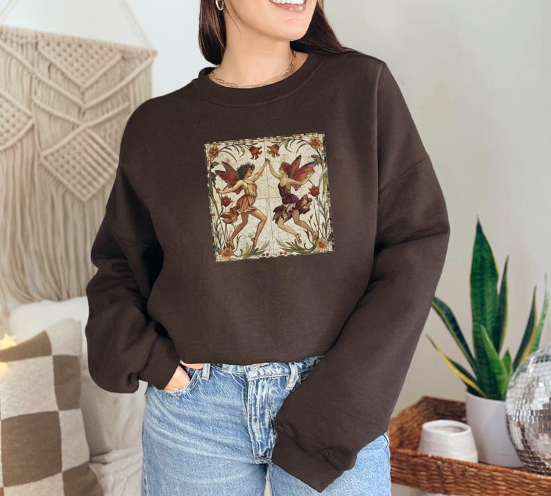 May include: A brown sweatshirt with a vintage-style illustration of two fairies dancing in a floral frame. The fairies are wearing colorful dresses and have wings.