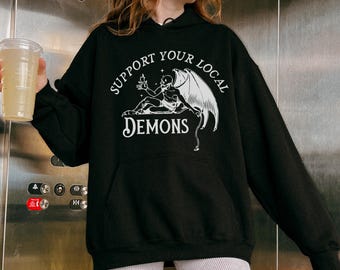 Peut inclure: Un sweat-shirt à capuche noir avec une impression graphique blanche représentant un démon ailé et le texte Support Your Local Demons. Le vêtement comprend une poche kangourou à l'avant, des manches longues et une capuche à cordon de serrage.