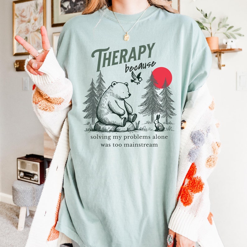 Color: Bay/Light Blue
Text: Therapy because solving my problems alone was too mainstream
Image Graphic: Bear sitting on along, rabbit sitting on ground beside bear, pine trees, bird, and red sun in background, peaceful outdoors design