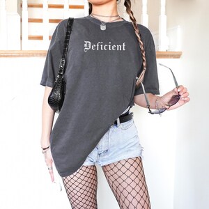 May include: A dark gray oversized t-shirt with the word "Deficient" printed in white on the front. The shirt is worn with light blue denim shorts and black fishnet stockings.