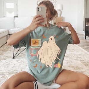 Comfort Colors 1717 Shirt
Color: Bay (light green blue)
Graphic: Two cartoon-style ghosts watch a witch flying on a TV screen surrounded by stars, popcorn, VHS tapes, and crescent moons.