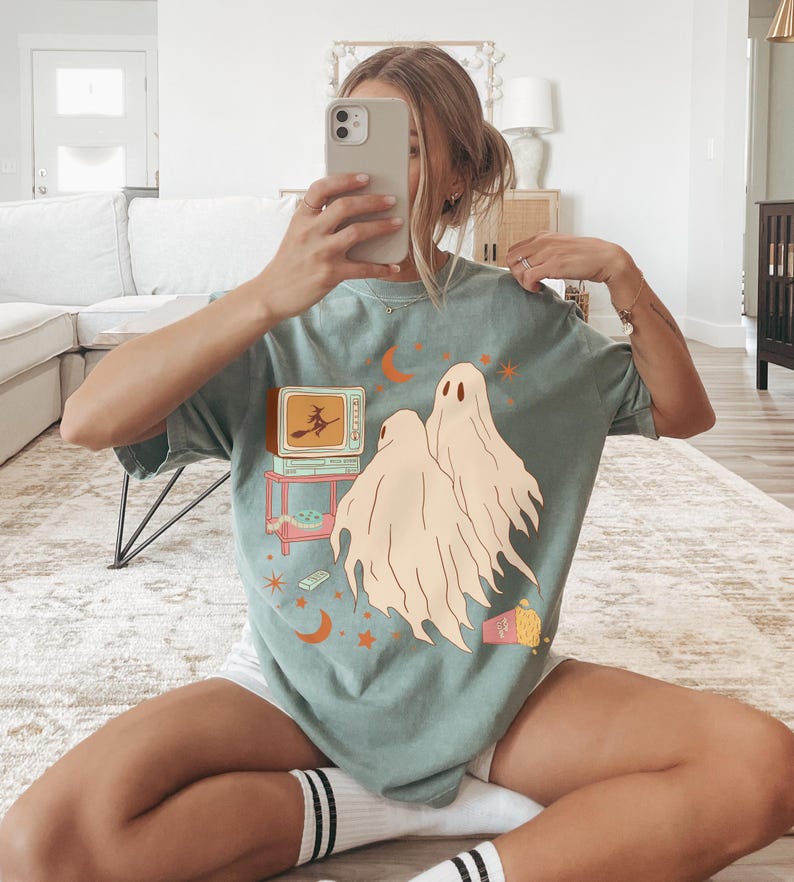 Comfort Colors 1717 Shirt
Color: Bay (light green blue)
Graphic: Two cartoon-style ghosts watch a witch flying on a TV screen surrounded by stars, popcorn, VHS tapes, and crescent moons.