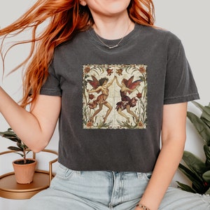 Pu&ograve; includere: Una maglietta corta grigia scura con un'illustrazione in stile vintage di due fate con le ali, circondate da fiori e foglie. Le fate indossano abiti colorati e si tengono per mano.