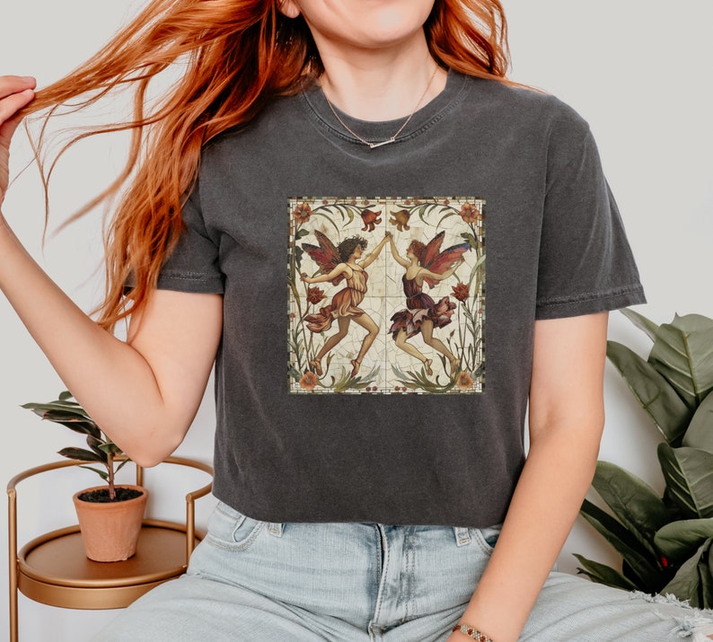 Pu&ograve; includere: Una maglietta corta grigia scura con un'illustrazione in stile vintage di due fate con le ali, circondate da fiori e foglie. Le fate indossano abiti colorati e si tengono per mano.