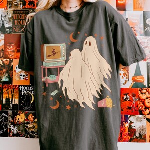 Comfort Colors 1717 Shirt
Color: Pepper (textured gray)
Graphic: Two cartoon-style ghosts watch a witch flying on a TV screen surrounded by stars, popcorn, VHS tapes, and crescent moons.
