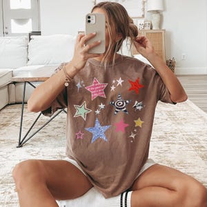 Comfort Colors 1717 T-Shirt
Color: Espresso Brown
Graphic: Colorful collage-style star graphic featuring various hand-drawn and patterned stars scattered across the chest. The stars include a mix of animal prints, stripes, solid colors, and textures, with some metallic and doodle-like accents.