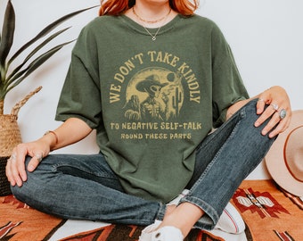 May include: A green t-shirt with a graphic of an alien wearing a cowboy hat and smoking a cigarette. The text on the shirt reads "We don't take kindly to negative self-talk around these parts."
