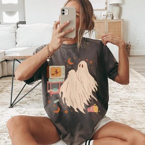 Comfort Colors 1717 Shirt
Color: Pepper (textured gray)
Graphic: Two cartoon-style ghosts watch a witch flying on a TV screen surrounded by stars, popcorn, VHS tapes, and crescent moons.