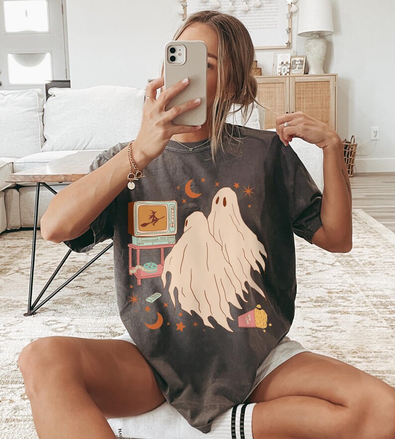 Comfort Colors 1717 Shirt
Color: Pepper (textured gray)
Graphic: Two cartoon-style ghosts watch a witch flying on a TV screen surrounded by stars, popcorn, VHS tapes, and crescent moons.