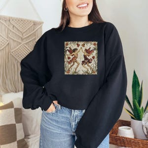 May include: Black sweatshirt with a vintage illustration of two fairies dancing in a floral frame. The fairies are wearing red and white dresses with wings.