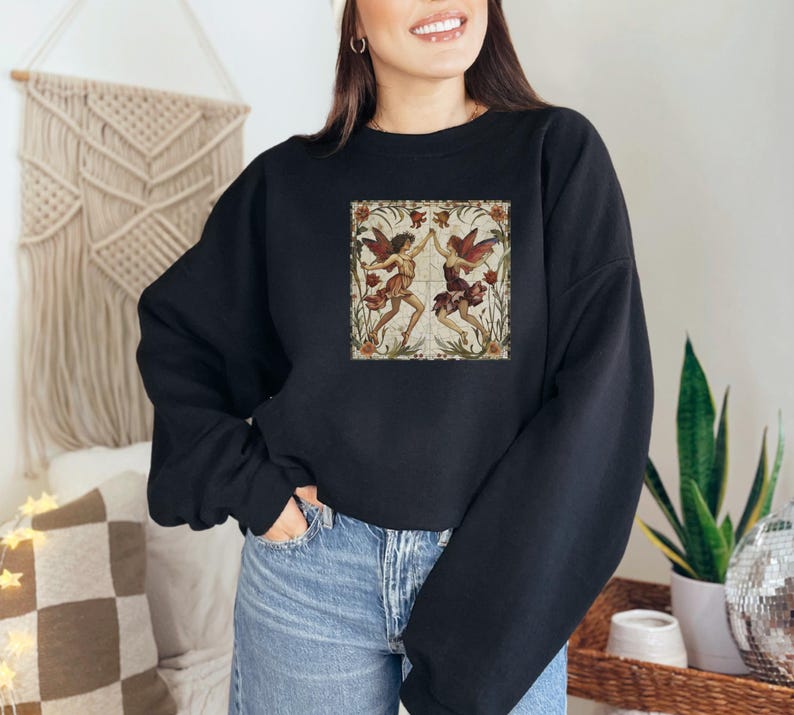 May include: Black sweatshirt with a vintage illustration of two fairies dancing in a floral frame. The fairies are wearing red and white dresses with wings.