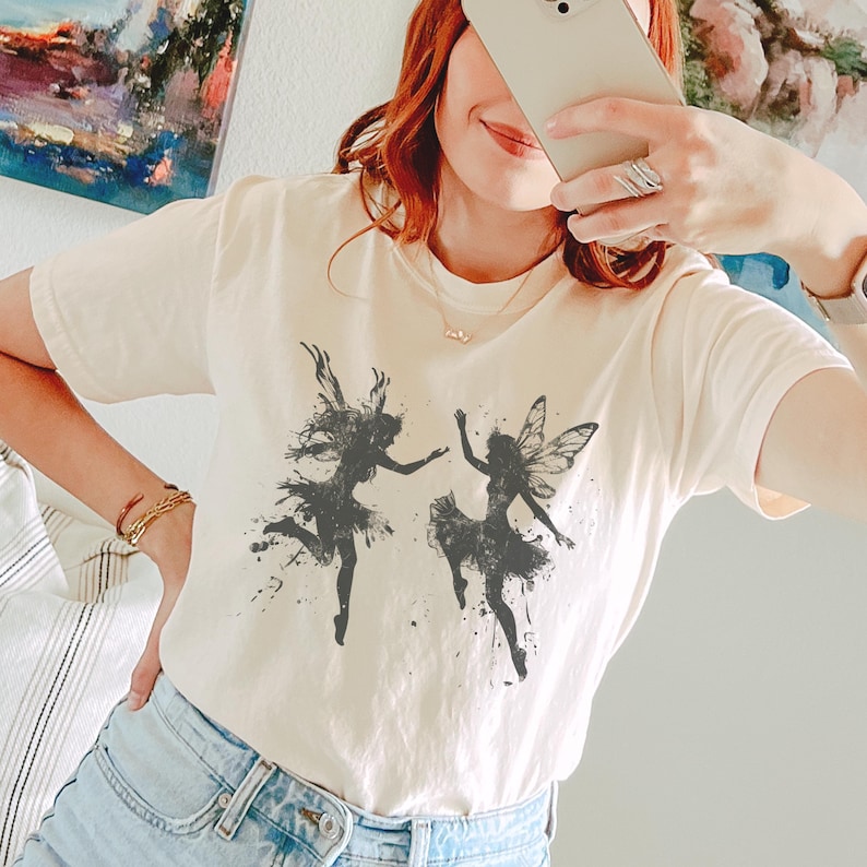 May include: A white t-shirt with a black and white graphic of two fairies dancing. The fairies are silhouetted against a white background with black splatters of paint.