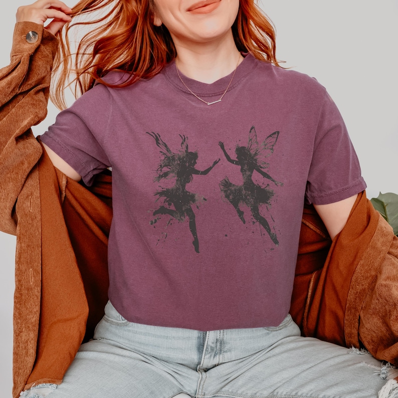 May include: A purple t-shirt with a graphic of two fairies dancing. The fairies are black silhouettes against a white background.