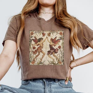 Pu&ograve; includere: Una maglietta marrone con un'illustrazione in stile vintage di due fate che ballano in una cornice floreale. Le fate indossano abiti colorati e hanno le ali. La cornice &egrave; realizzata con un motivo a mosaico.