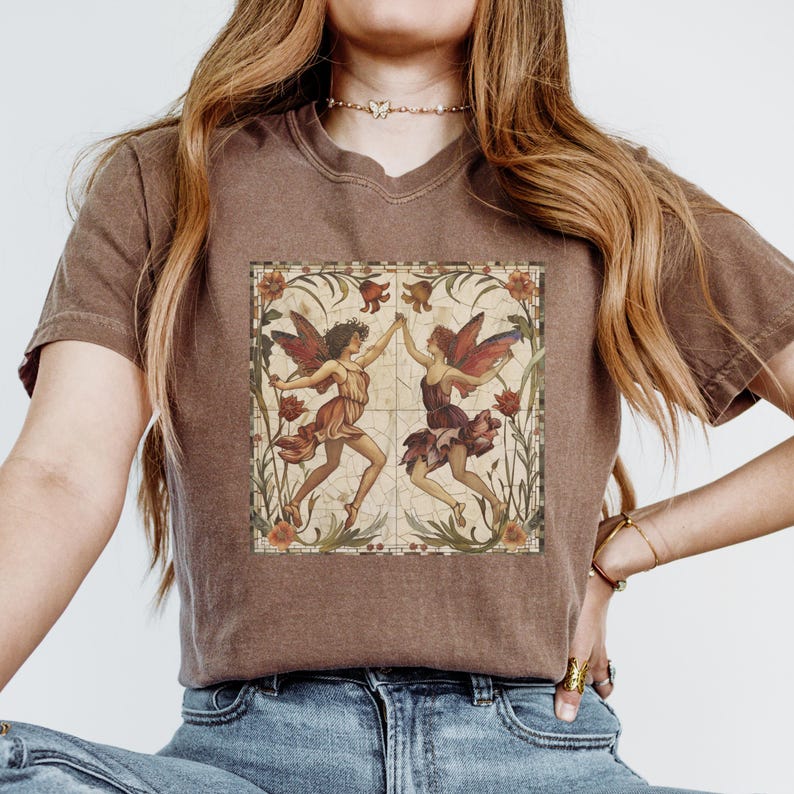 Pu&ograve; includere: Una maglietta marrone con un'illustrazione in stile vintage di due fate che ballano in una cornice floreale. Le fate indossano abiti colorati e hanno le ali. La cornice &egrave; realizzata con un motivo a mosaico.