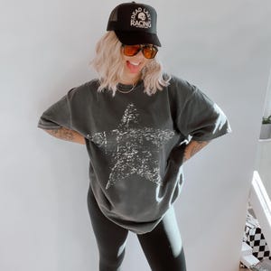 May include: A black trucker hat with the text "DEAD LAST RACING" and a skull graphic. The person is wearing a dark gray oversized t-shirt with a large white star graphic and black leggings.