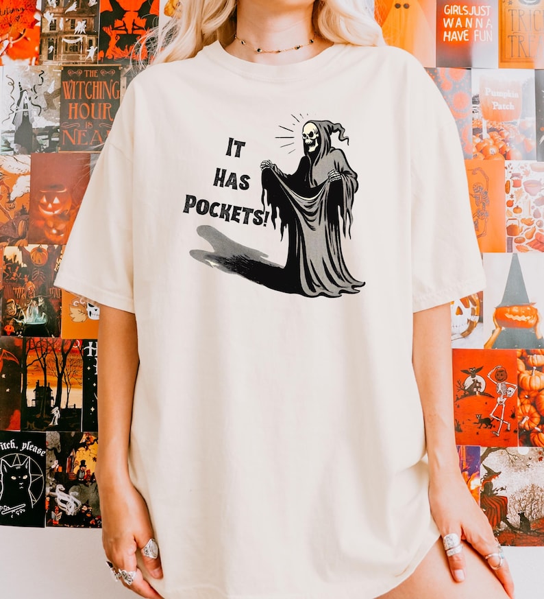 Op de afbeelding: Een wit T-shirt met een afbeelding van een skelet dat een gewaad draagt en het openhoudt om zakken te laten zien. De tekst "IT HAS POCKETS!" staat onder de afbeelding.
