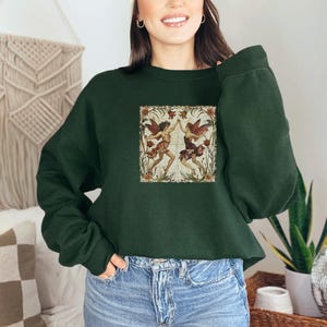 May include: A dark green cropped sweatshirt with a vintage illustration of two fairies dancing in a floral setting. The fairies are wearing flowing dresses and have wings. The illustration is surrounded by a decorative border.