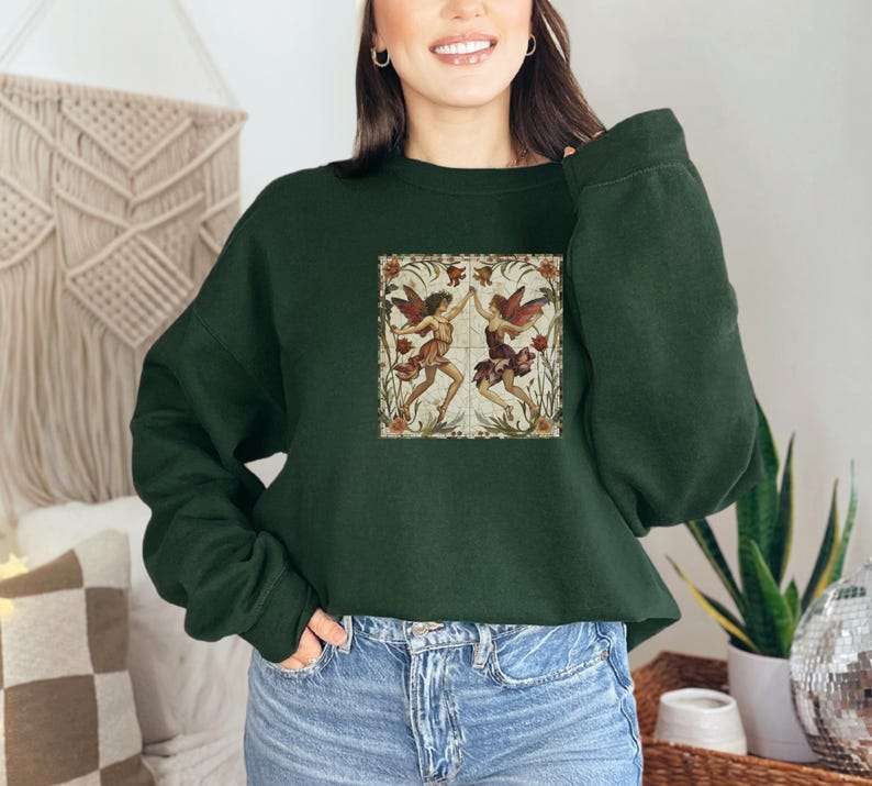 May include: A dark green cropped sweatshirt with a vintage illustration of two fairies dancing in a floral setting. The fairies are wearing flowing dresses and have wings. The illustration is surrounded by a decorative border.