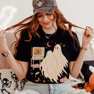 Comfort Colors 1717 Shirt
Color: Black
Graphic: Two cartoon-style ghosts watch a witch flying on a TV screen surrounded by stars, popcorn, VHS tapes, and crescent moons.