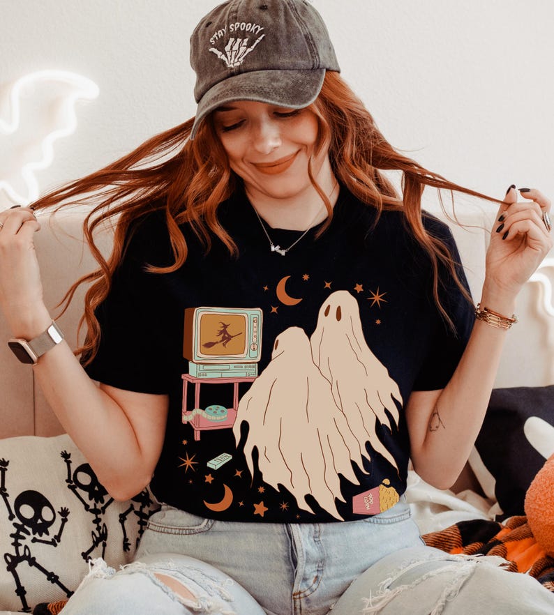 Comfort Colors 1717 Shirt
Color: Black
Graphic: Two cartoon-style ghosts watch a witch flying on a TV screen surrounded by stars, popcorn, VHS tapes, and crescent moons.