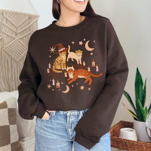 May include: A dark brown sweatshirt featuring a whimsical illustration of three cats adorned with witch hats, crowns, and surrounded by candles, moons, and stars. The cats are in shades of orange, tan, and white.