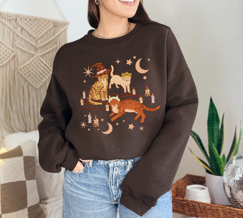 May include: A dark brown sweatshirt featuring a whimsical illustration of three cats adorned with witch hats, crowns, and surrounded by candles, moons, and stars. The cats are in shades of orange, tan, and white.