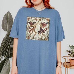 Pu&ograve; includere: Una maglietta oversize blu con un'illustrazione vintage di due fate che ballano in una cornice floreale.