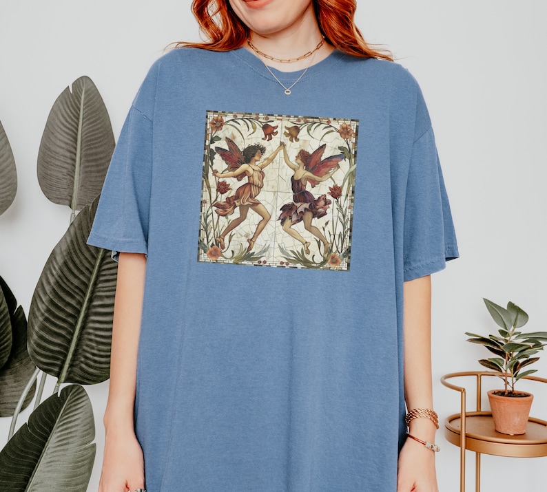 Pu&ograve; includere: Una maglietta oversize blu con un'illustrazione vintage di due fate che ballano in una cornice floreale.