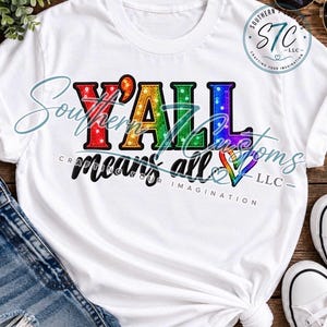 May include: A white t-shirt featuring the text Y'ALL in large, multi-colored, sequin-style letters with a rainbow gradient. Below, the words means all are printed in black script, accompanied by a small, multi-colored heart outline.