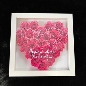 May include: A framed artwork featuring a heart-shaped arrangement of pink paper roses. A white outline of the United States is superimposed on the roses. The phrase "Home is where the heart is" is written in white.
