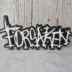 May include: A black and white sign featuring the word "FORSAKEN" in a graffiti-style font. The white text has a black outline. The sign is set against a grey surface, with a wooden backdrop.