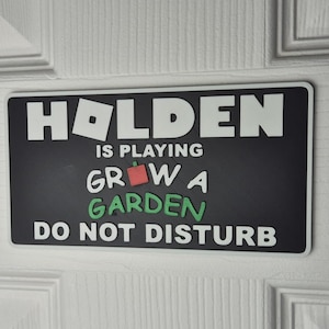 Puede incluir: Un letrero rectangular negro con letras blancas y verdes que dice "HOLDEN IS PLAYING GROW A GARDEN DO NOT DISTURB". El letrero tiene un borde blanco y está montado en una puerta blanca.