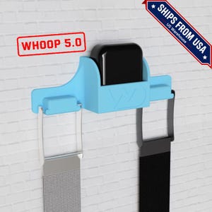May include: A light blue plastic charging dock for a Whoop 5.0 fitness tracker. The dock features a central black charging unit and two side slots holding a silver metallic mesh band and a black fabric band, with Whoop 5.0 text visible.
