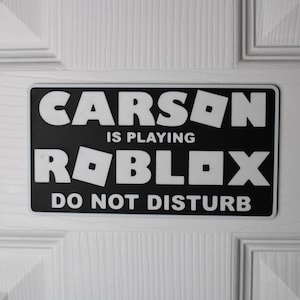 Peut inclure: Panneau rectangulaire noir et blanc fixé sur une porte blanche. Le panneau indique "CARSON IS PLAYING ROBLOX DO NOT DISTURB" en gras, police sans-serif. Le logo Roblox est intégré au texte.