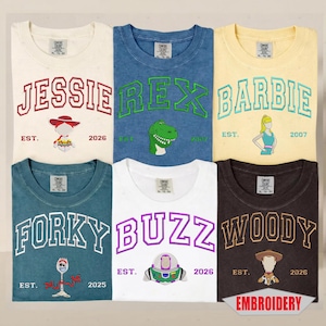 May include: Six short-sleeve t-shirts in various colors, each featuring embroidered character names and graphics. The shirts display Jessie, Rex, Barbie, Forky, Buzz, and Woody with established dates. Each shirt has a Comfort Colors brand neck label.