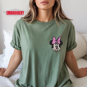 May include: Olive green t-shirt featuring an embroidered Minnie Mouse design with a pink bow. The word "EMBROIDERY" is printed in a red banner in the upper left corner. The shirt has rolled-up sleeves and a relaxed fit.