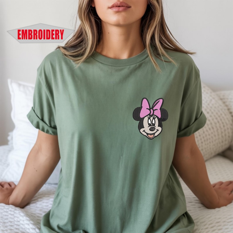 May include: Olive green t-shirt featuring an embroidered Minnie Mouse design with a pink bow. The word "EMBROIDERY" is printed in a red banner in the upper left corner. The shirt has rolled-up sleeves and a relaxed fit.
