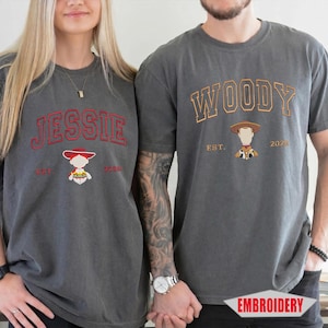 May include: Two charcoal gray t-shirts featuring embroidered designs. The left shirt displays the name JESSIE in red with a character graphic and EST. 2025. The right shirt displays the name WOODY in orange with a character graphic and EST. 2025.