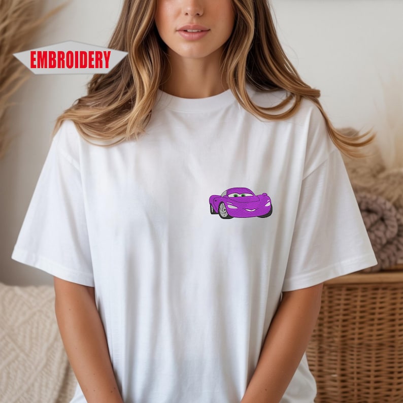 May include: White t-shirt with a small, embroidered purple cartoon car on the chest. The car has a smiling face and silver wheels. The word "EMBROIDERY" is printed in red on the top left of the image.