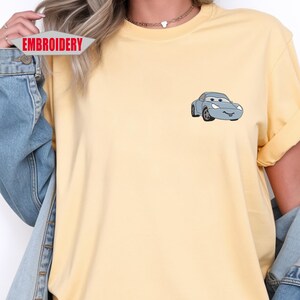May include: A light yellow t-shirt featuring a small, light blue cartoon car graphic on the chest. The word "EMBROIDERY" is printed on a white and red banner. The shirt has rolled-up sleeves.