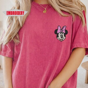 May include: A magenta-colored t-shirt with an embroidered Minnie Mouse design on the chest. The design features Minnie's face with a pink bow. The word "EMBROIDERY" is printed on a white banner in the upper left corner.