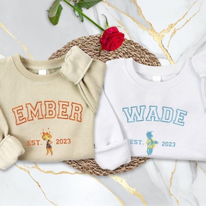 May include: Two crewneck sweatshirts, one tan and one white. The tan sweatshirt has "EMBER" in orange lettering and a small graphic. The white sweatshirt has "WADE" in blue lettering and a graphic. Both say "EST. 2023".